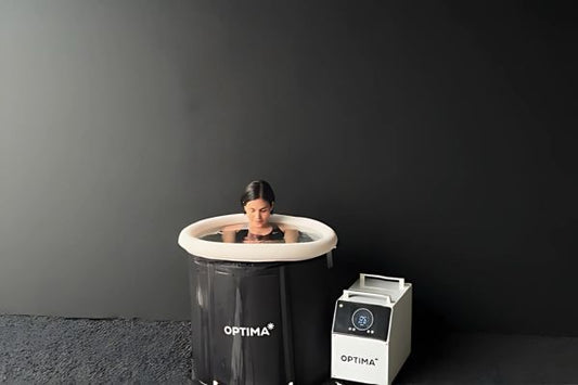 Woman fully immersed in OPTIMA cold plunge tub next to chiller unit, black background.